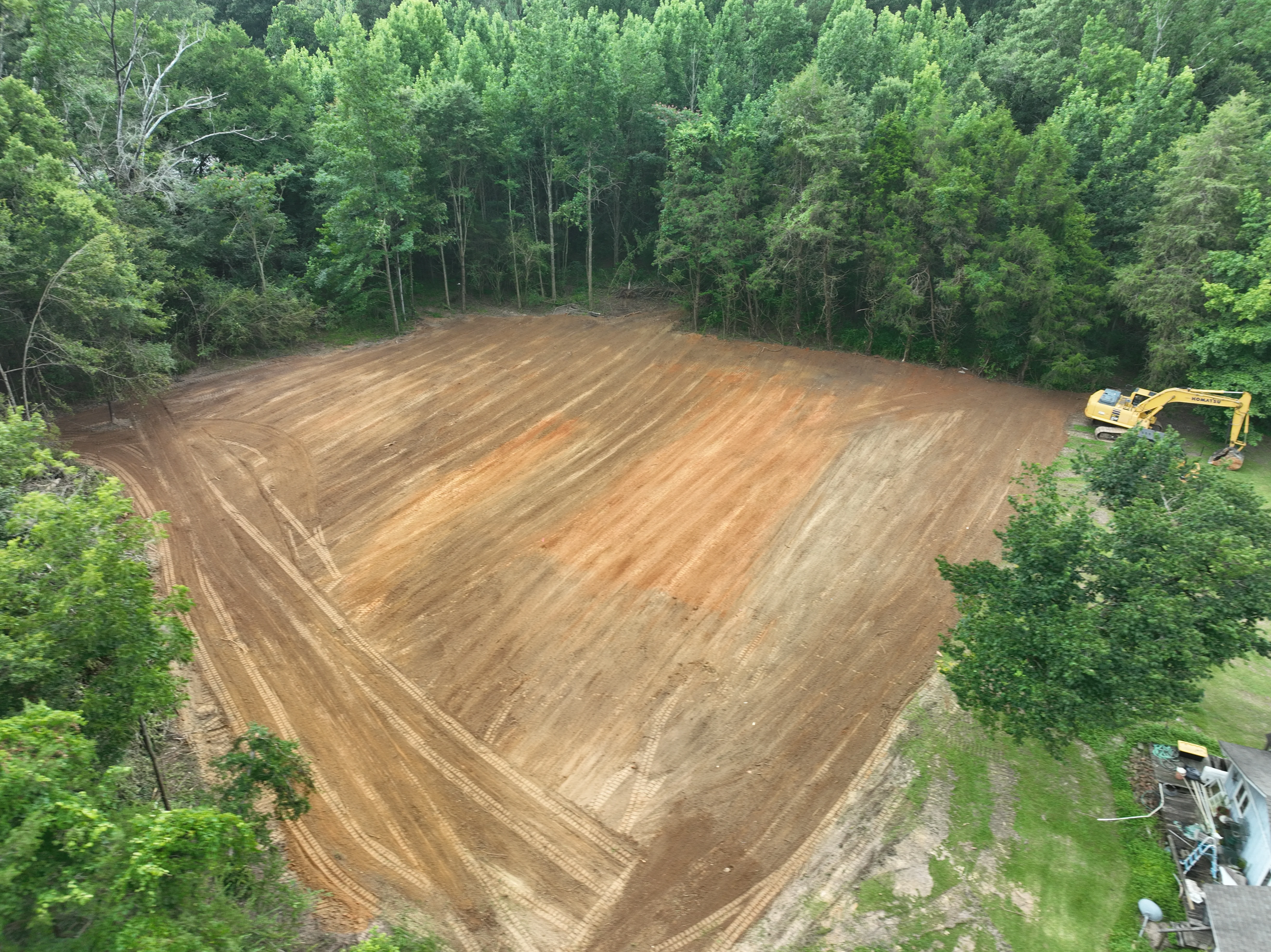 Aerial view of cleared wooded lot with excavator and graded brown soil surrounded by green forest